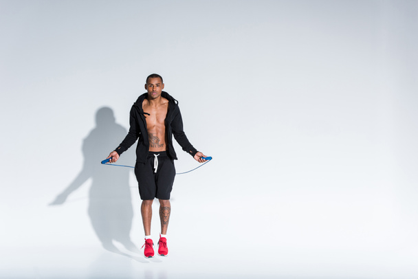 young african american sportsman exercising with skipping rope and looking at camera on grey - Foto, imagen