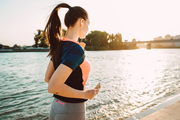 jovem menina fitness de cabelos longos em roupas esportivas corre pelo rio, ao sol, ouvindo música em fones de ouvido
 - Foto, Imagem
