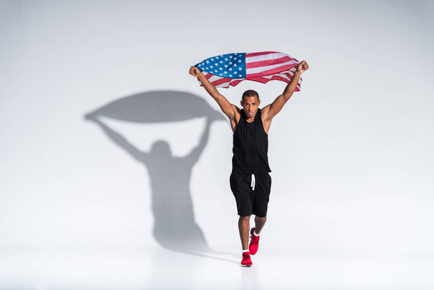 jonge Afro-Amerikaanse sportman met Amerikaanse vlag camera kijken op grijs - Foto, afbeelding