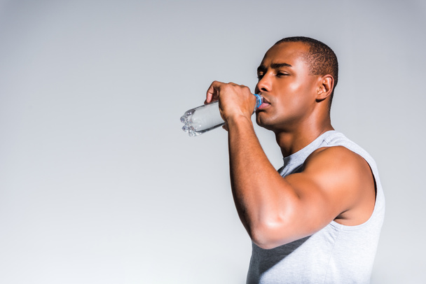 молодой африканский американский спортсмен пьет воду из бутылки, изолированной на сером
 - Фото, изображение