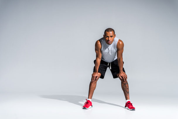 vista completa de atlético joven afroamericano deportista de pie con las manos en las rodillas y mirando a la cámara en gris
 - Foto, Imagen