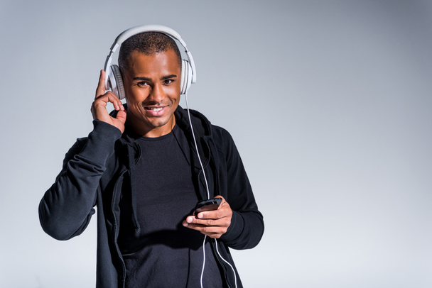 молодий афроамериканський чоловік в навушниках використовує смартфон і посміхається на камеру ізольовано на сірому
  - Фото, зображення