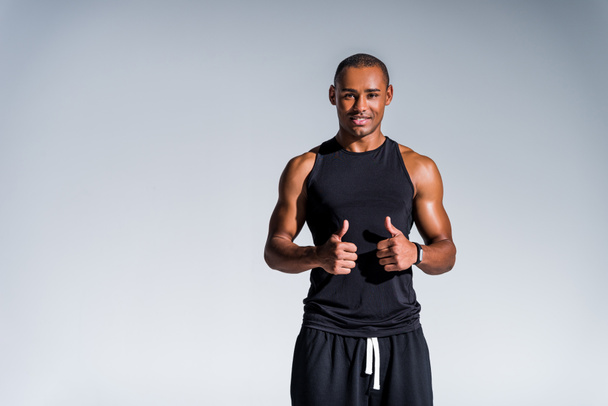 feliz esportista afro-americano mostrando polegares para cima e sorrindo para a câmera isolada em cinza
 - Foto, Imagem