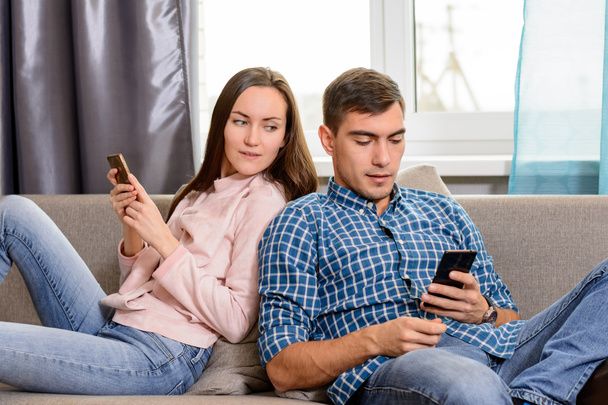 Νεαρό ζευγάρι κάθεται στον καναπέ και χρησιμοποιώντας το smartphone, το κορίτσι spies μέσω ένας τύπος κινητό τηλέφωνο - Φωτογραφία, εικόνα