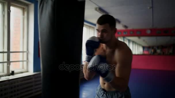 Мускулистый мужчина профессиональный боксер тренируется, пробивая грушу в тренажерном зале в боксёрских перчатках с голыми грудями
. - Кадры, видео