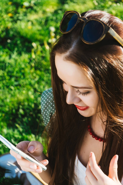 Vrouw zoek iets in mobiel, SMS, verslaafd, zit op het gras in het park - Foto, afbeelding