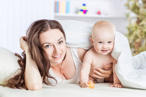 concept of happy mothers: happy mother and her year-old child playing on the sofa in the bedroom - Photo, Image