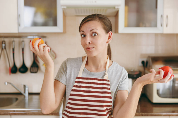 Una joven atractiva mujer pensativa en un delantal decide elegir un tomate rojo o amarillo en la cocina. Concepto de dieta. Estilo de vida saludable. Cocinar en casa. Preparar alimentos
. - Foto, Imagen