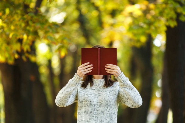 Piękny szczęśliwy uśmiechający się Brunet Kobieta w białym swetrze, chowając się za czerwone książki w Upadek miasta park na ciepły dzień. Autumn golden odchodzi. Koncepcja czytania. - Zdjęcie, obraz