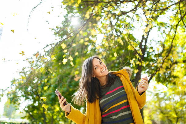 Güzel bir mutlu neşeli kahverengi saçlı kadın sarı ceket ve çizgili longsleeve sonbahar Şehir Parkı cep telefonunda sıcak bir günde konuşmak. Altın yaprak. - Fotoğraf, Görsel