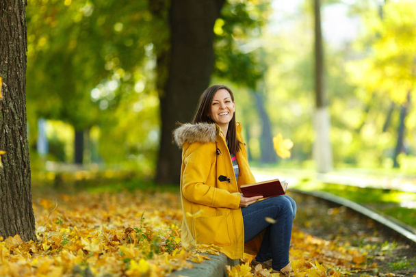 Piękne kobiety Brunet szczęśliwy w żółtym fartuchu i dżinsy samodzielnie znajduje się w parku w pobliżu torów tramwajowych i czyta książkę w ciepły dzień jesieni. Żółty jesienny liść. - Zdjęcie, obraz
