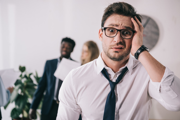 bewildered young businessman touching head and looking at camera at office - Foto, Imagem