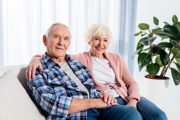 portrait of smiling senior wife and husband looking at camera while resting on sofa at home - Foto, Bild