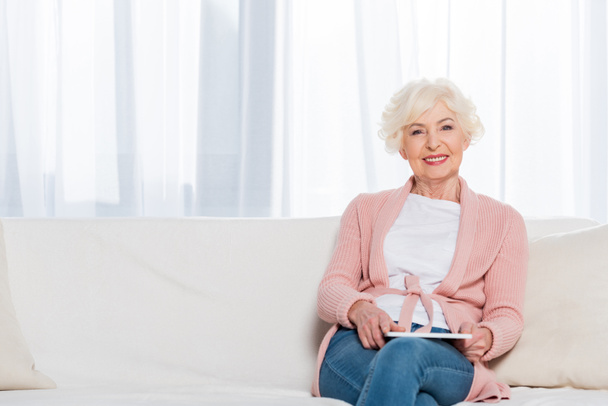 portrait of smiling senior woman with tablet in hands resting on sofa and looking at camera at home - Foto, afbeelding