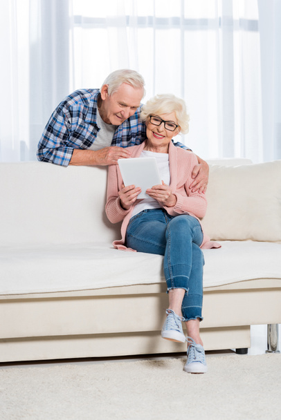 senior man hugging wife with digital tablet in hands on sofa at home - Foto, Imagen