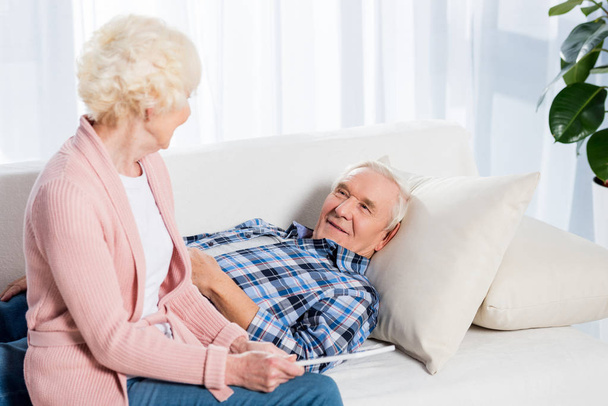 smiling senior man looking at wife with tablet in hands while resting on sofa at home - Foto, Imagen