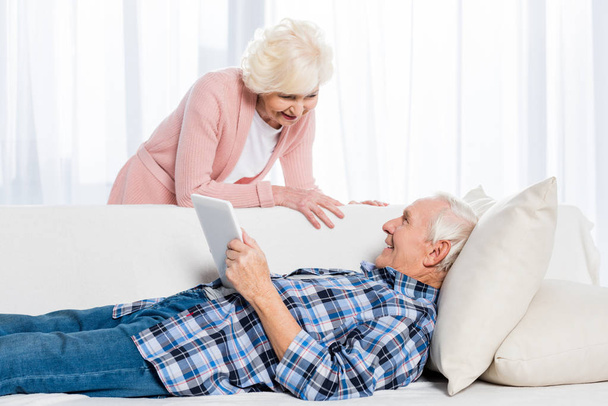 homem sênior com tablet olhando para a esposa sorridente por perto, enquanto descansa no sofá em casa
 - Foto, Imagem