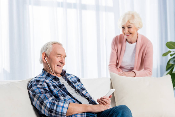 selektywne focus starszy człowiek w słuchawki do słuchania muzyki, podczas gdy żona stojących w pobliżu w domu - Zdjęcie, obraz