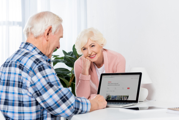 portrét usměvavá starší ženy při pohledu na muže pomocí přenosného počítače s airbnb logem doma - Fotografie, Obrázek