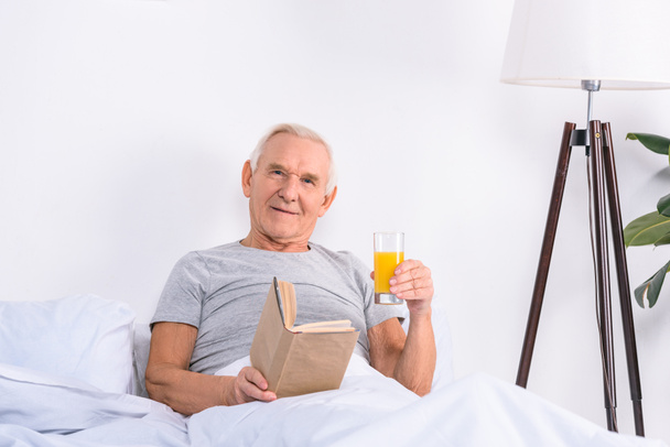 Porträt eines älteren Mannes mit einem Glas Saft und einem Buch in der Hand, der zu Hause im Bett in die Kamera blickt - Foto, Bild