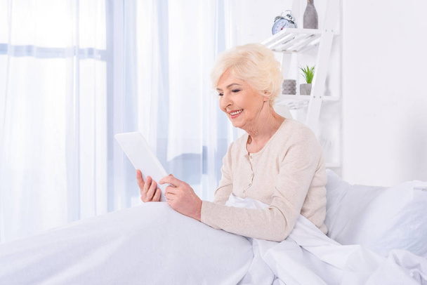 side view of happy senior woman with tablet lying in bed at home - Foto, afbeelding