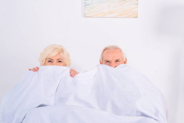 obscured view of senior wife and husband covering faces with blanket and looking at camera - Fotó, kép