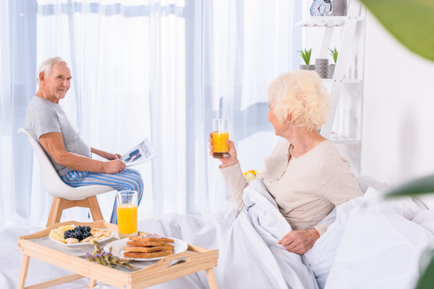 vista laterale della donna anziana che fa colazione a letto mentre il marito con il giornale seduto a tavola in camera da letto
 - Foto, immagini