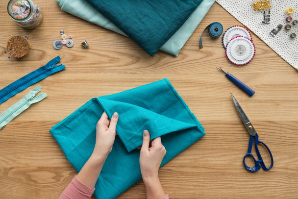 vista superior de las manos femeninas recortadas tela plegable en el lugar de trabajo costurera con tijeras y botones
 - Foto, Imagen