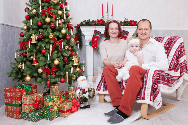Christmas or New year celebration. Portrait of cheerful young family of three people near the Christmas tree with xmas gifts. A fireplace with christmas stocking on background. Happy holidays - 写真・画像