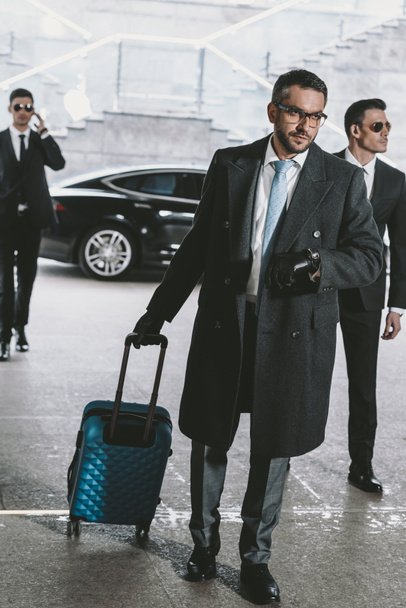 handsome businessman walking with travel bag from car and looking at watch - Foto, imagen