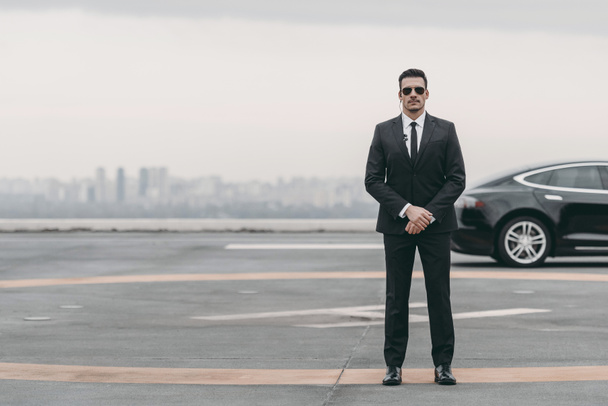 serious bodyguard standing with sunglasses and security earpiece on helipad and looking at camera - Photo, Image