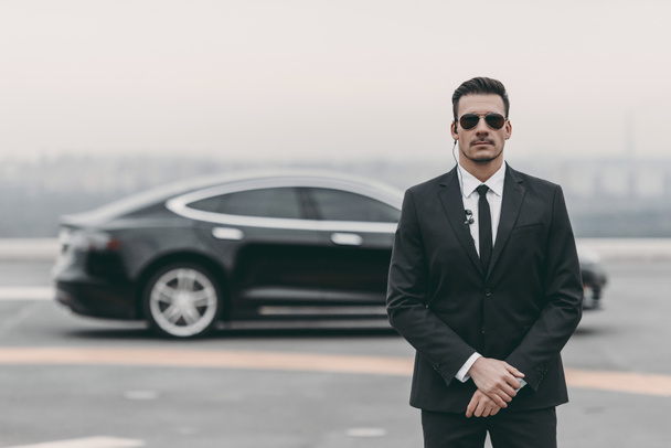 serious bodyguard standing with sunglasses and security earpiece on helipad  - Photo, Image