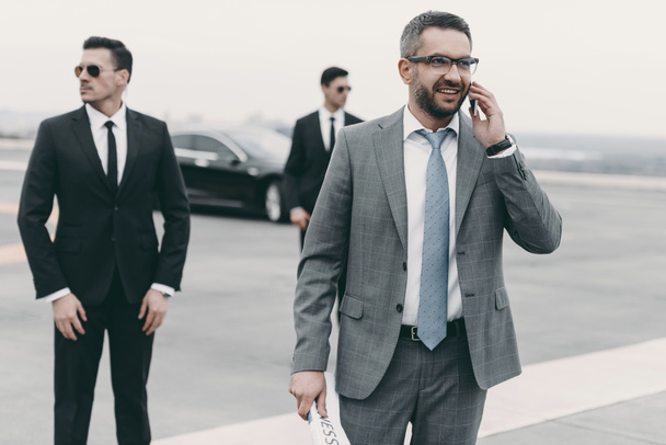 smiling businessman walking with bodyguards and talking by smartphone  - Photo, Image
