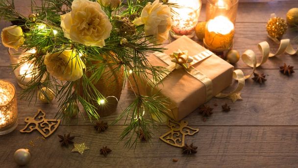Decoración de Navidad. Regalo, velas, luces, bolas doradas sobre una mesa rústica de madera. Composición de ramas de pino y rosas inglesas en un jarrón
. - Foto, Imagen