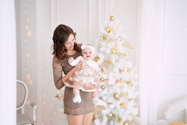 Φόρεμα κορίτσι μωρό φοράει χαριτωμένος και στήριγμα κεφαλής με τη μητέρα, κοντά στο χριστουγεννιάτικο δέντρο σε εορταστικά διακοσμημένο δωμάτιο με γιρλάντα από τα φώτα. Με έκπληξη ρολόγια στη φωτογραφική μηχανή, με φόντο μια σειρά από φωτεινό φωτιά - Φωτογραφία, εικόνα
