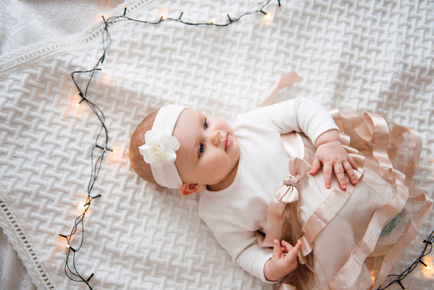 Meisje van de baby dragen schattige jurk en hoofdband, ligt op een witte cover in de feestelijk versierde kamer met garland lichten. Met verbazing horloges in de camera, op een achtergrond een aantal felle branden. Warm worden - Foto, afbeelding