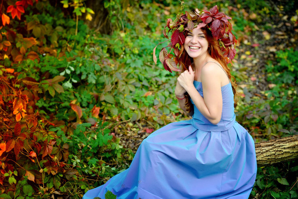 Charming girl in a wreath from autumn leaves, in a blue dress sitting on a log on the background of red and green bushes in a Sunny day and cheerfully laughs - Fotoğraf, Görsel