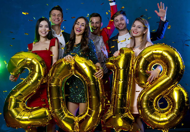 The year 2018 is coming! Group of cheerful young people in beautiful evening dresses and costumes carrying gold colored numbers and throwing confetti - Φωτογραφία, εικόνα