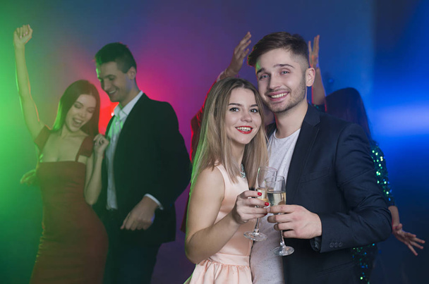 Ομάδα νέων χορεύουν χαρούμενος και ευτυχισμένος φίλους σε ένα πάρτι. Ένας άνδρας και μια γυναίκα χορεύει στο προσκήνιο και τσούγκριζαν τα ποτήρια με σαμπάνια - Φωτογραφία, εικόνα