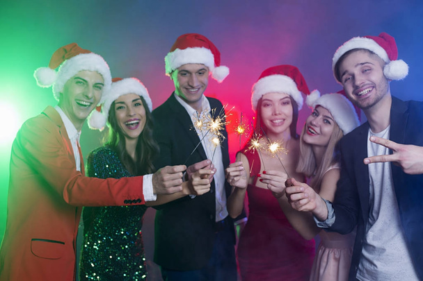 Happy new year and merry Christmas. Group of happy smiling friends in Santa hats with sparklers in their hands having fun on New Year's party. - Фото, зображення