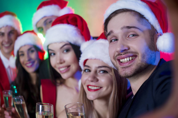 Group of smiling friends in Santa hats are photographed or make selfie on New Year's party - 写真・画像