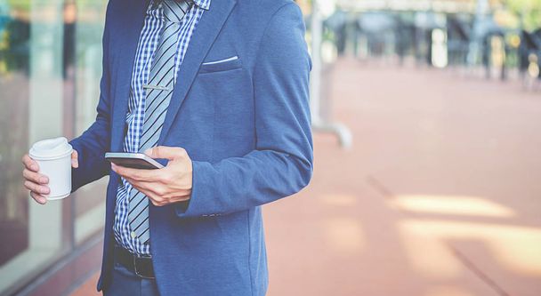 Handsome young businessman holding his laptop while using his phone and standing on the street - Photo, Image
