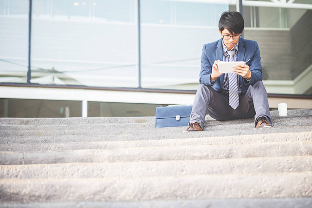 Красивый молодой бизнесмен, сидящий на лестнице и пользующийся смартфоном. Селективный фокус
. - Фото, изображение