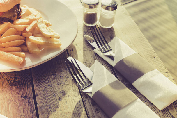 Hamburguesa, papas fritas y tenedores y cuchillos envueltos en una servilleta de papel servida en una mesa de madera, luz suave, filtro vintage
  - Foto, Imagen