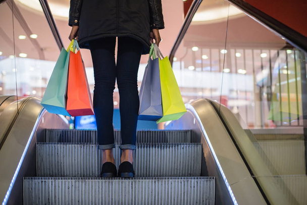 ショッピング モールのエスカレーターでカラフルなショッピング バッグを持つ女性の足 - 写真・画像
