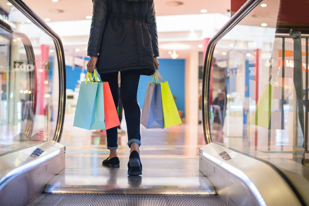 ショッピング モールのエスカレーターでカラフルなショッピング バッグを持つ女性の足 - 写真・画像