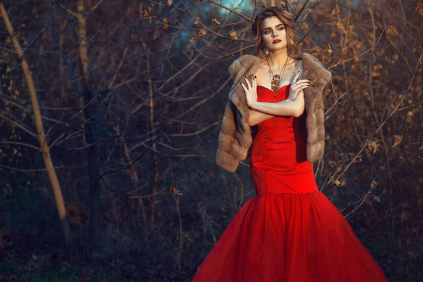 Gorgeous glam model with updo hair and beautiful make up wearing posh red fishtail dress and luxurious mink vest standing in dry autumn bushes with crossed arms - Photo, Image