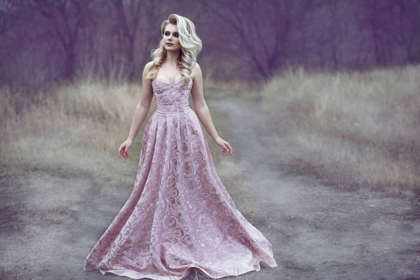 Retrato de una hermosa dama rubia con un exuberante peinado y hermoso maquillaje con lujoso vestido de bola de rosa de fresno brocado caminando por el estrecho camino en el bosque
 - Foto, Imagen