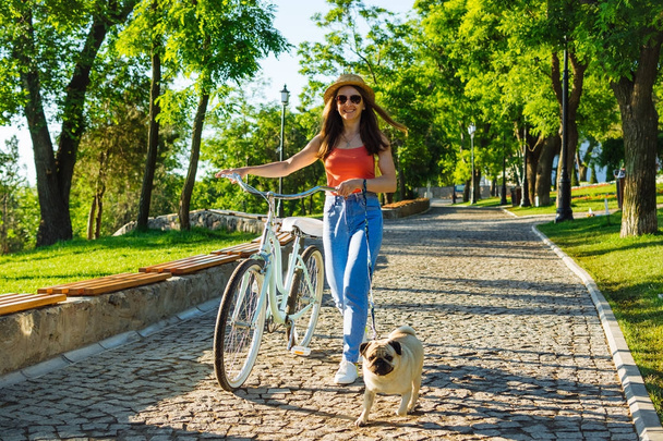 Young beautiful woman with vintage bicycle and pug walking in summer park. Green trees background     - Fotografie, Obrázek