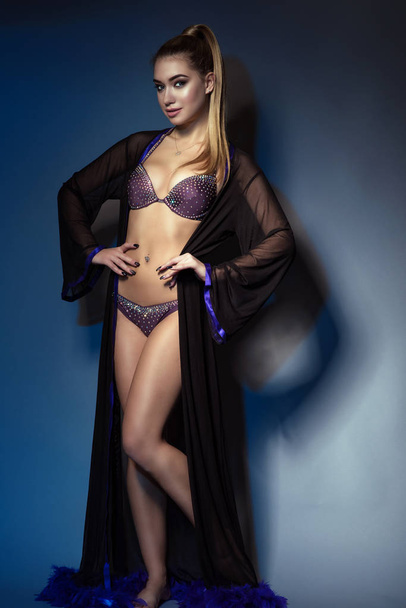 Full length portrait of young attractive blond model with high ponytail wearing beaded purple swimsuit and sheer black chiffon peignoir posing on blue background, her hands on hips - Fotografie, Obrázek